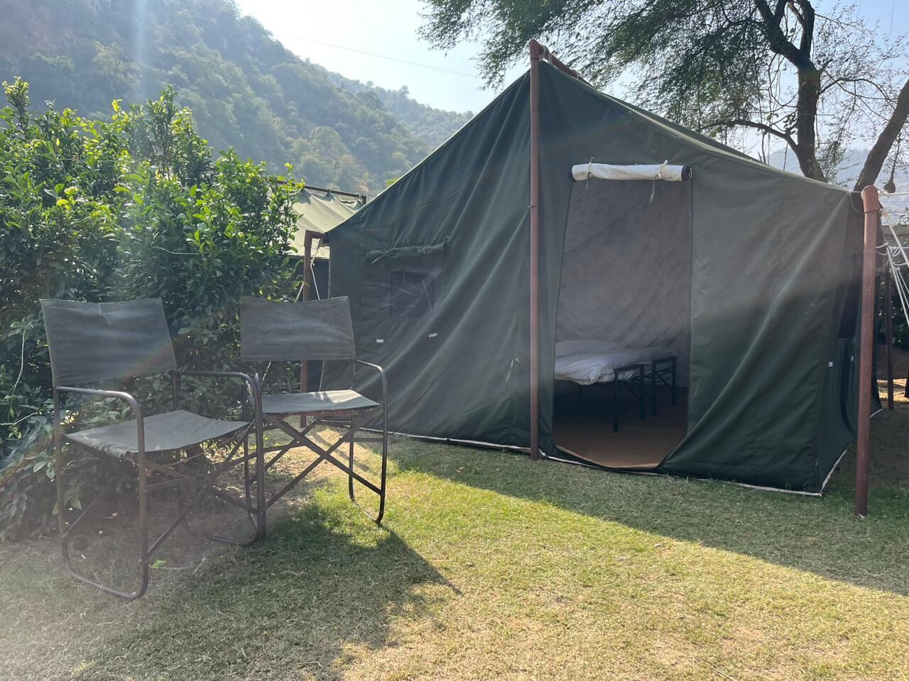 A tent with a bed inside, two folding chairs, and a grassy area with trees in the background.