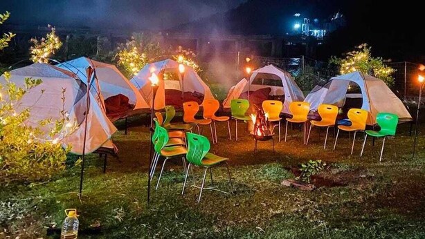 Une rangée de tentes, avec des chaises et un feu de camp, installée dans une zone herbeuse, avec des montagnes en arrière-plan.