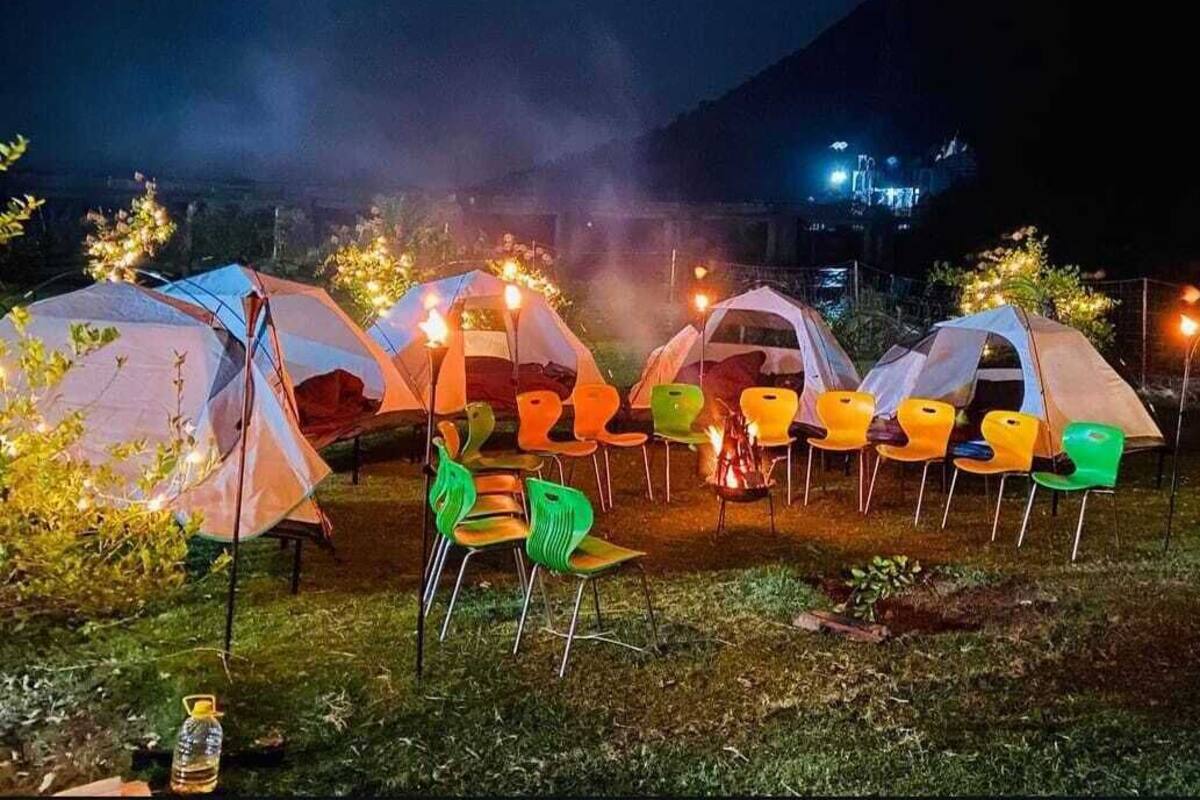A row of tents with chairs and a campfire, set in a grassy area with mountains in the background.