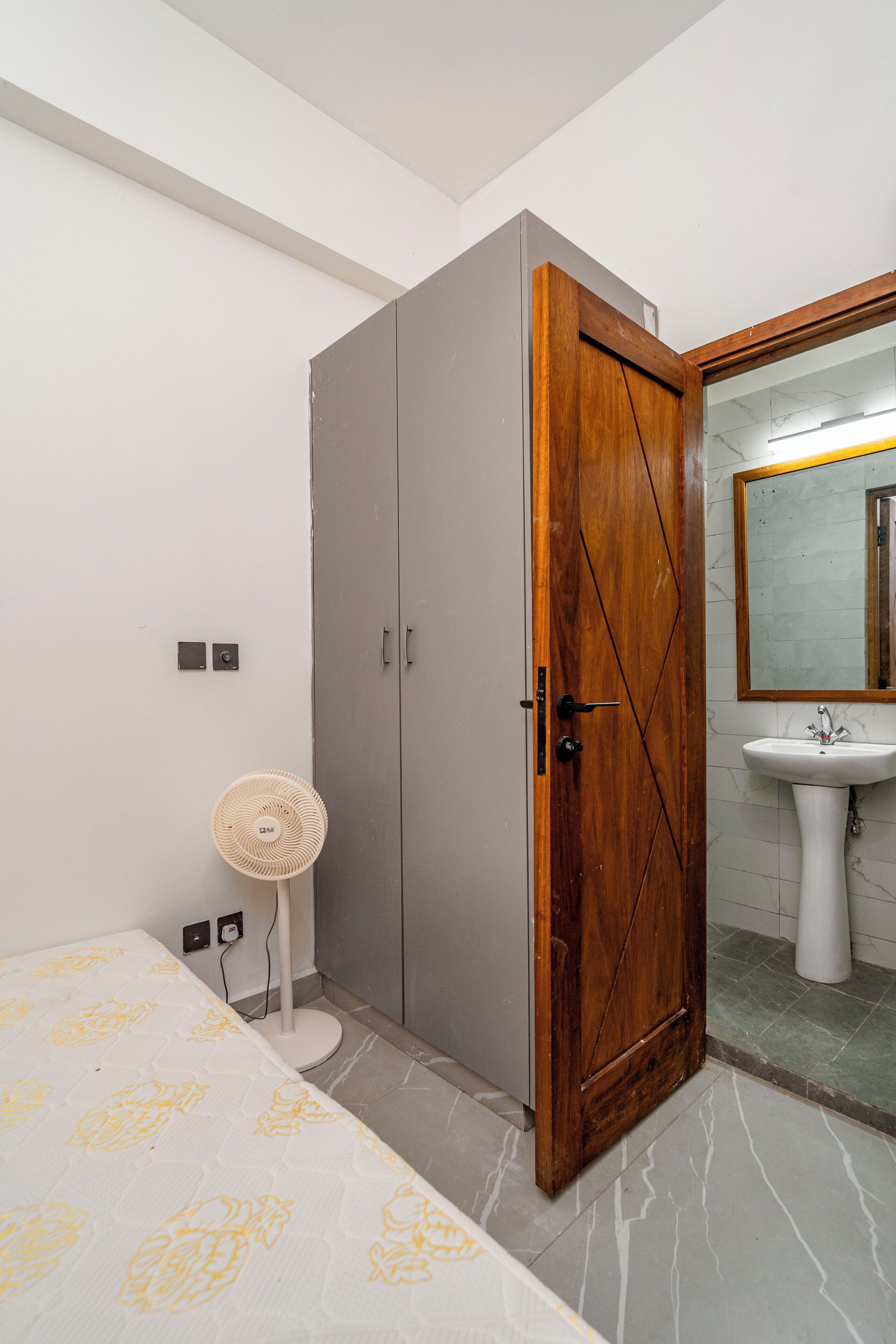 A bedroom with a bed, a wardrobe, a bedside fan, and a bathroom with a sink and mirror.
