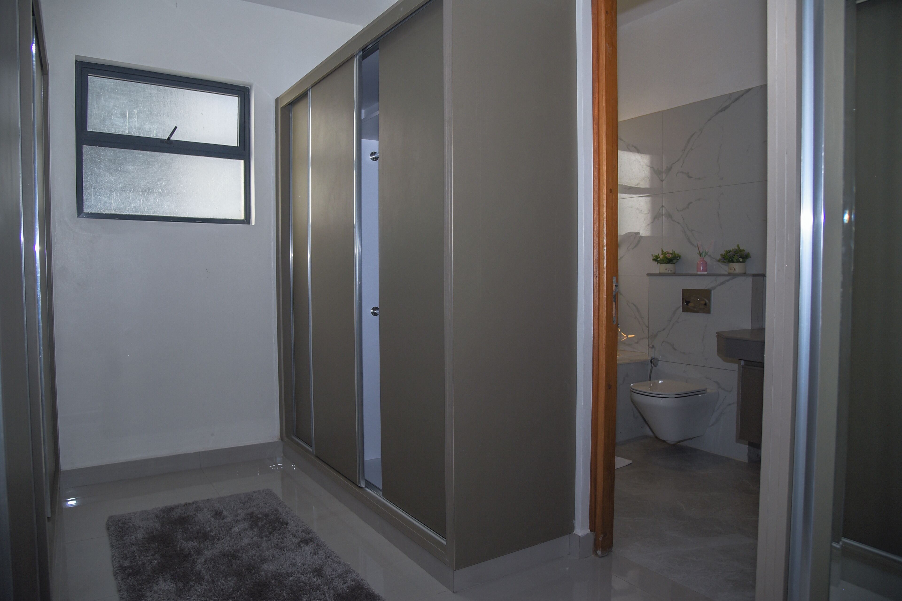 Ein Badezimmer mit Toilette, Waschbecken und Spiegel. Ein Fenster und eine Tür zu einem anderen Raum sind vorhanden.