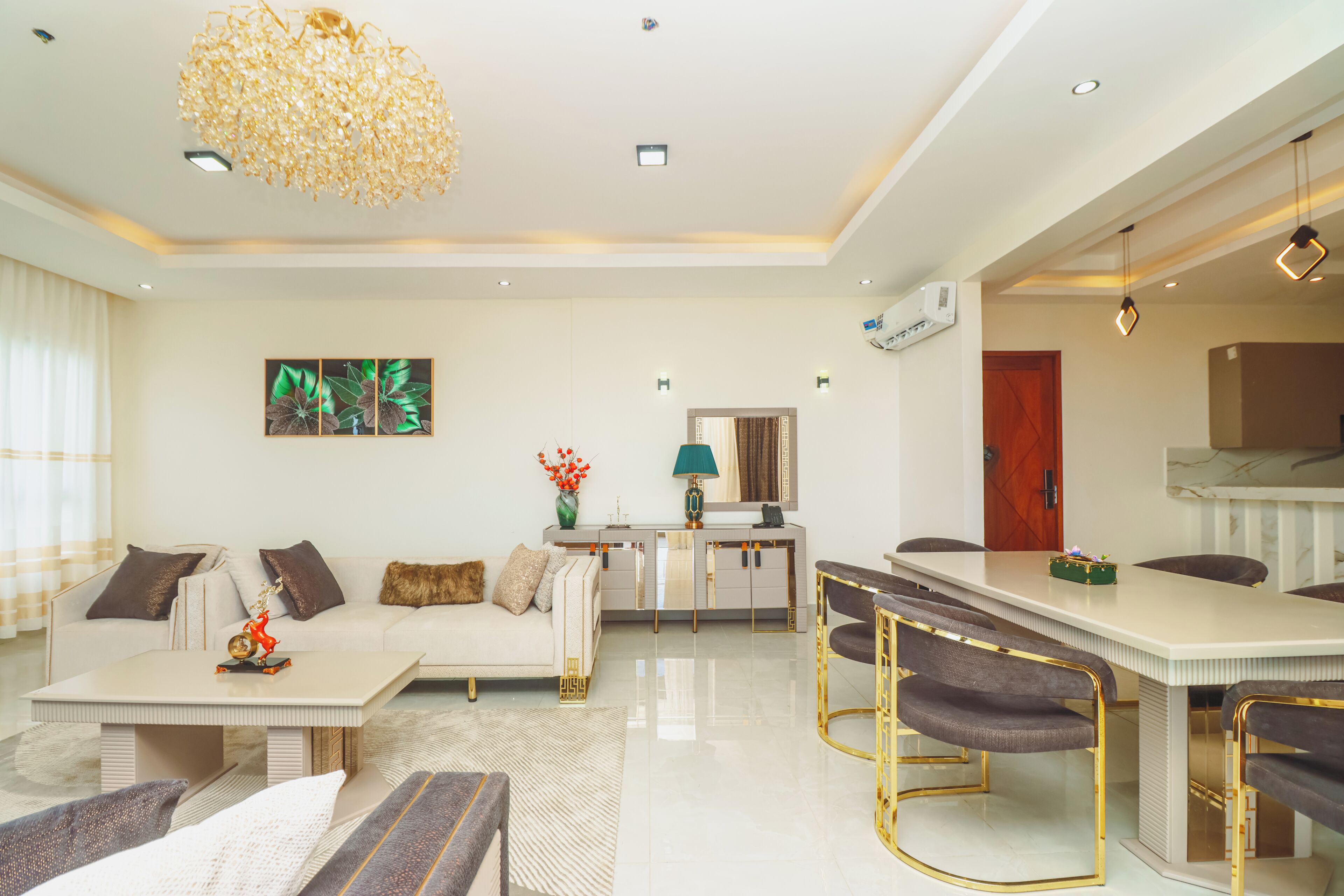 A modern living room with a sofa, coffee table, and dining area with chairs. There is a decorative chandelier, a painting on the wall, and a vase with flowers.