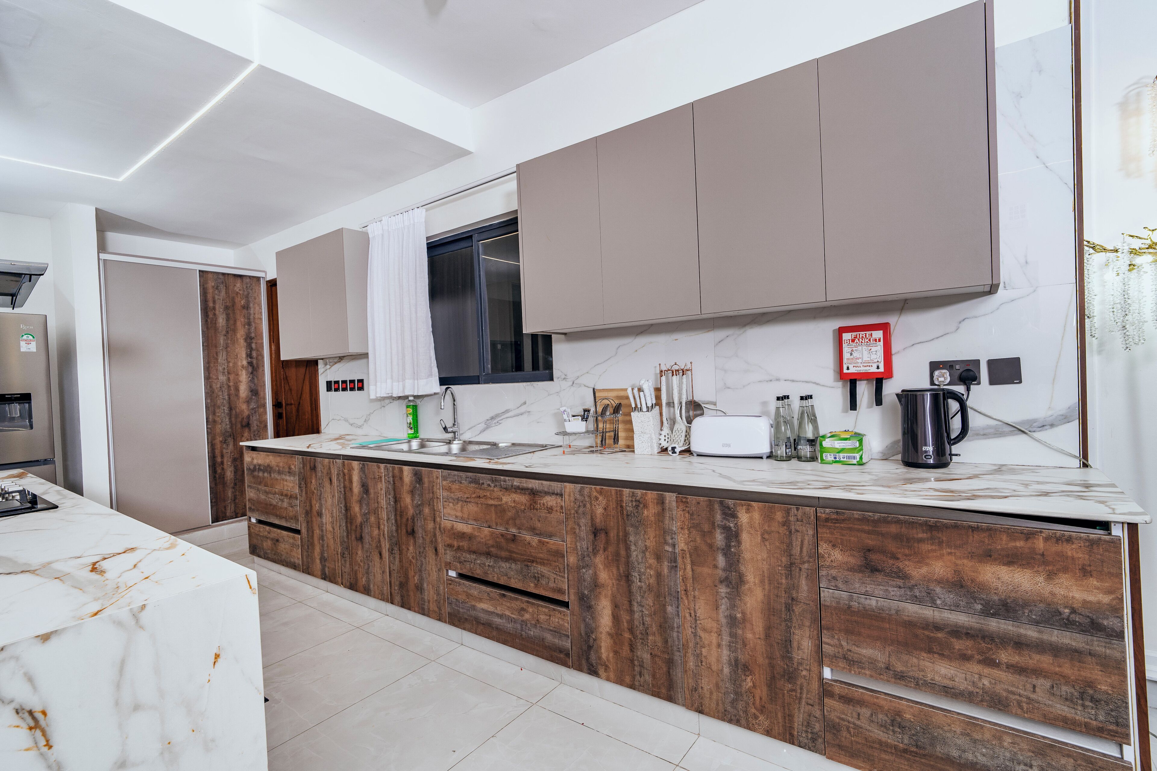 A modern kitchen with a marble countertop, wooden cabinets, a stainless steel sink, and a fire blanket mounted on the wall.