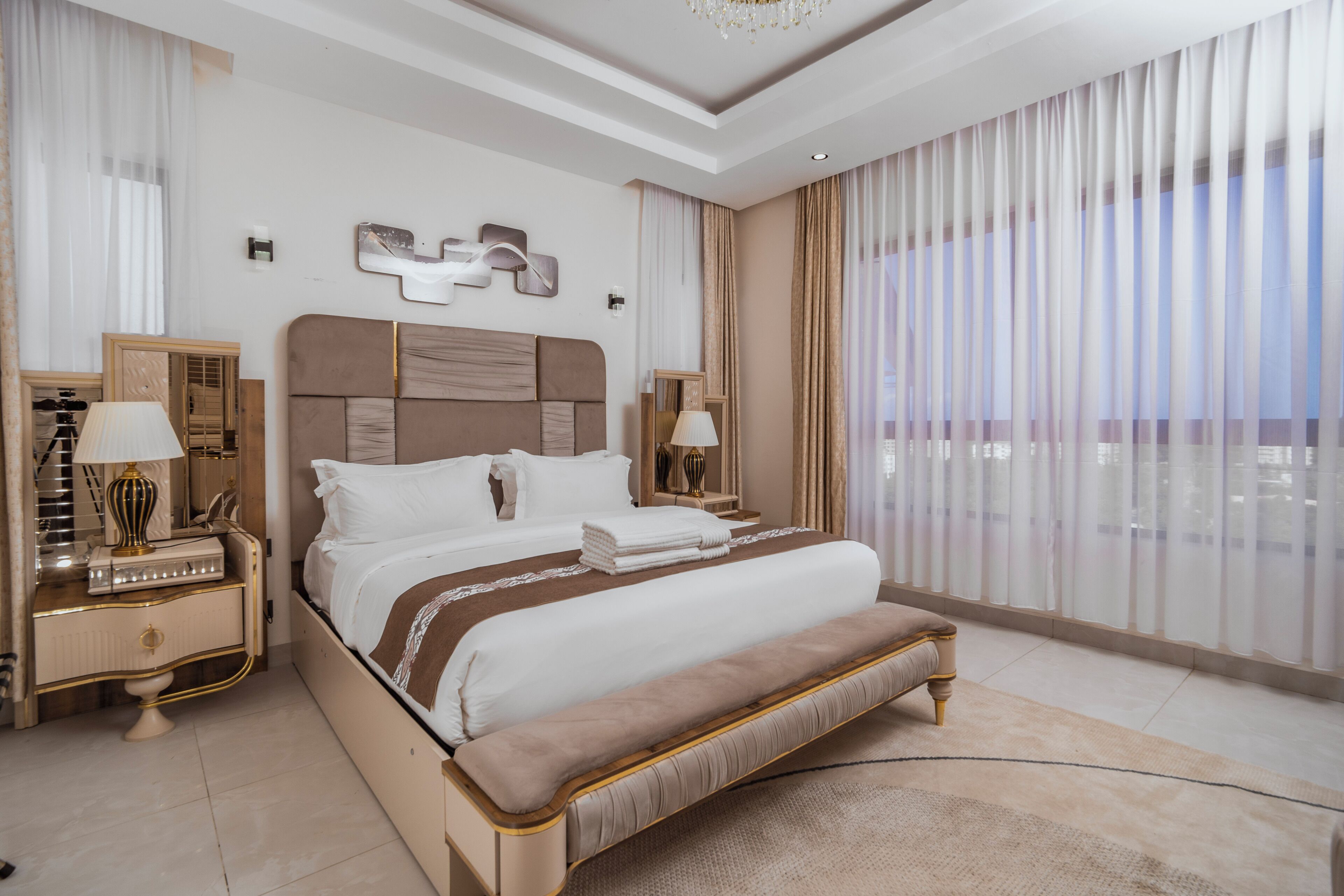 A spacious bedroom with a large bed, a vanity with a mirror, and a view of the city through sheer curtains.