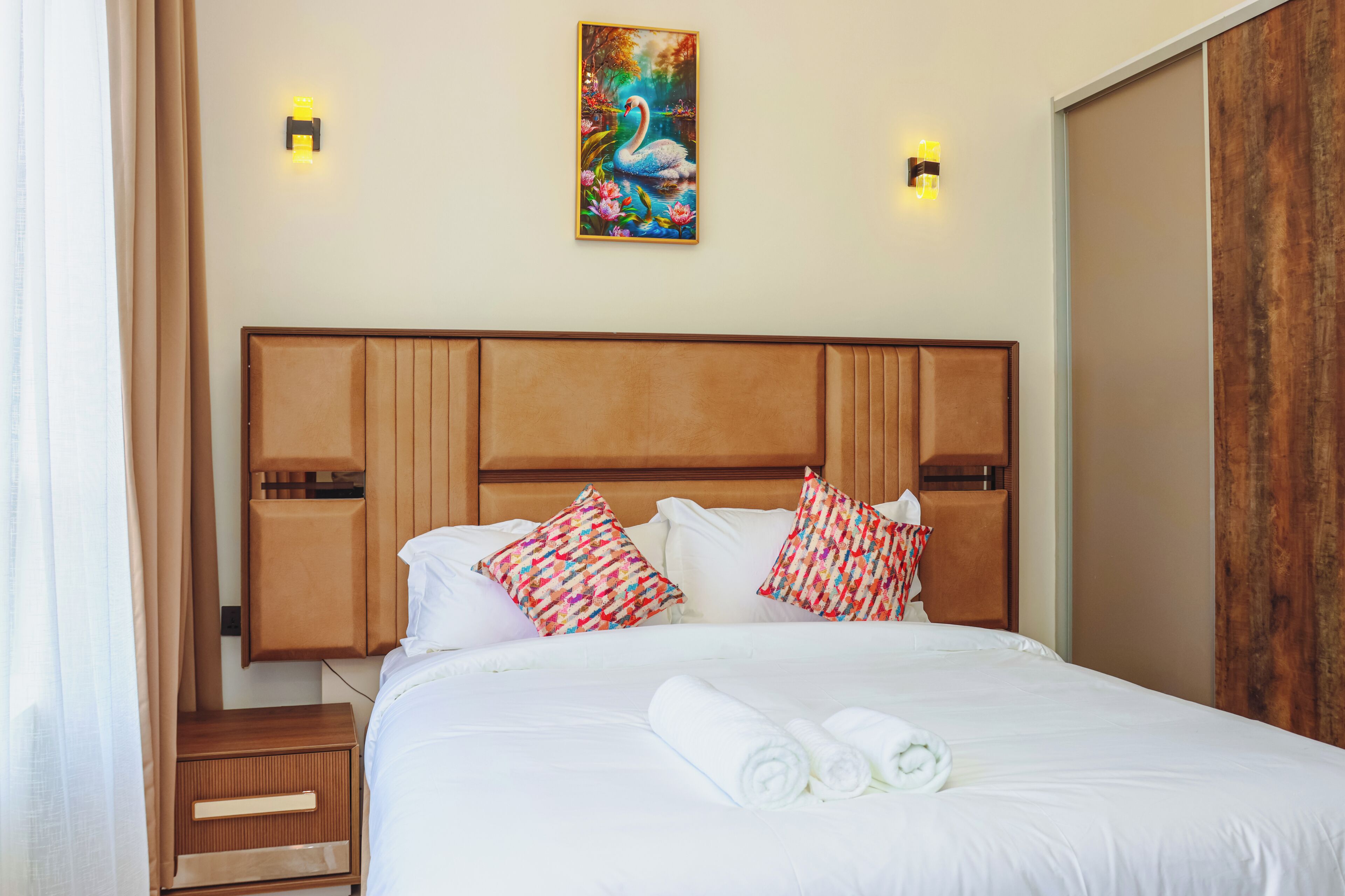 A hotel room with a wooden headboard, white bedding, decorative pillows, a painting on the wall, and two wall-mounted lights.