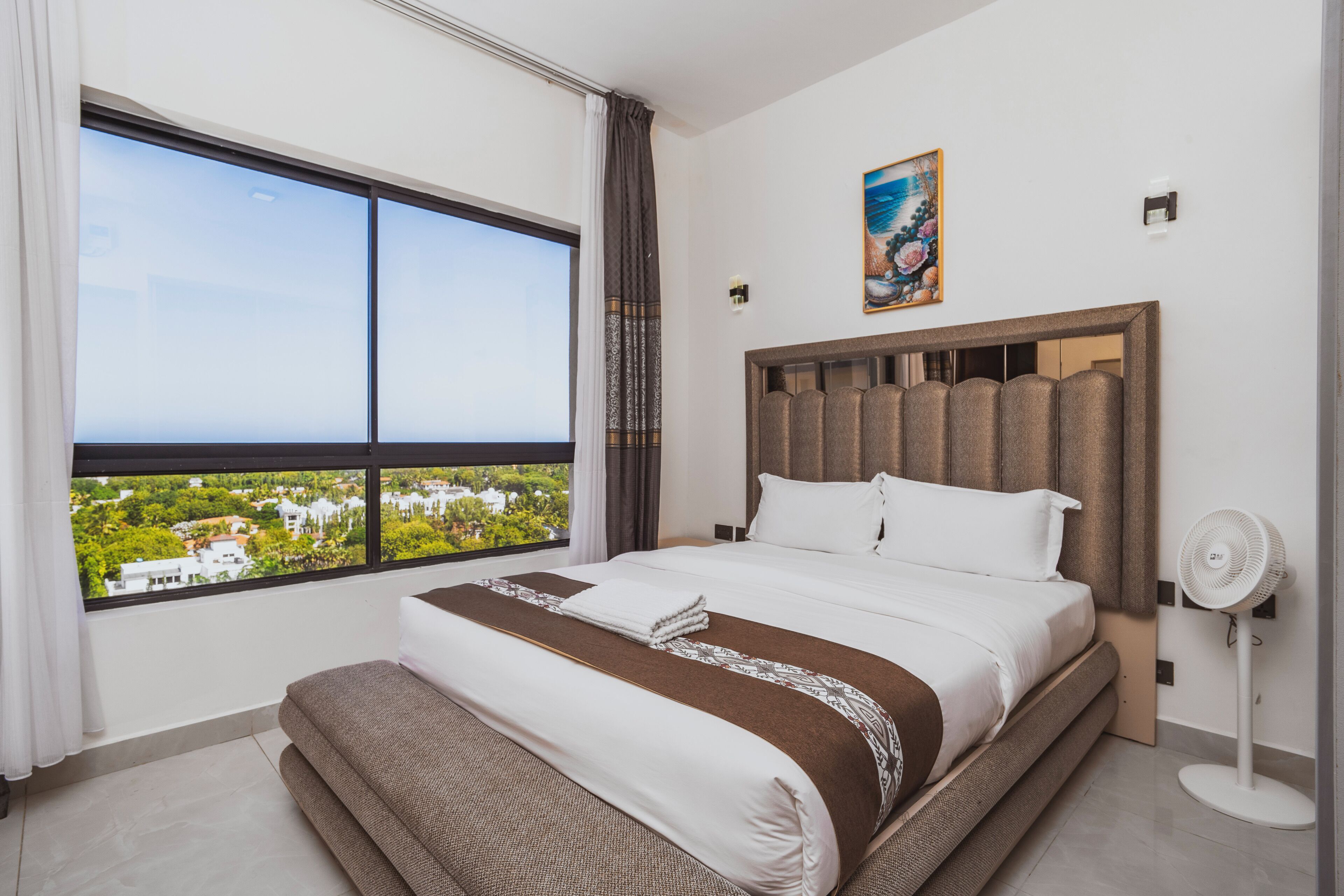A hotel room with a large window, a bed with a headboard, a bedside table, and a wall-mounted fan.