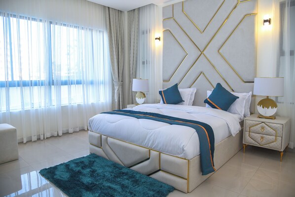 A modern bedroom with a large bed, two bedside tables, a nightstand, a fluffy rug, and a view of the outdoors through sheer curtains. - Mira Heights Suite & Conferencing (Mombasa)