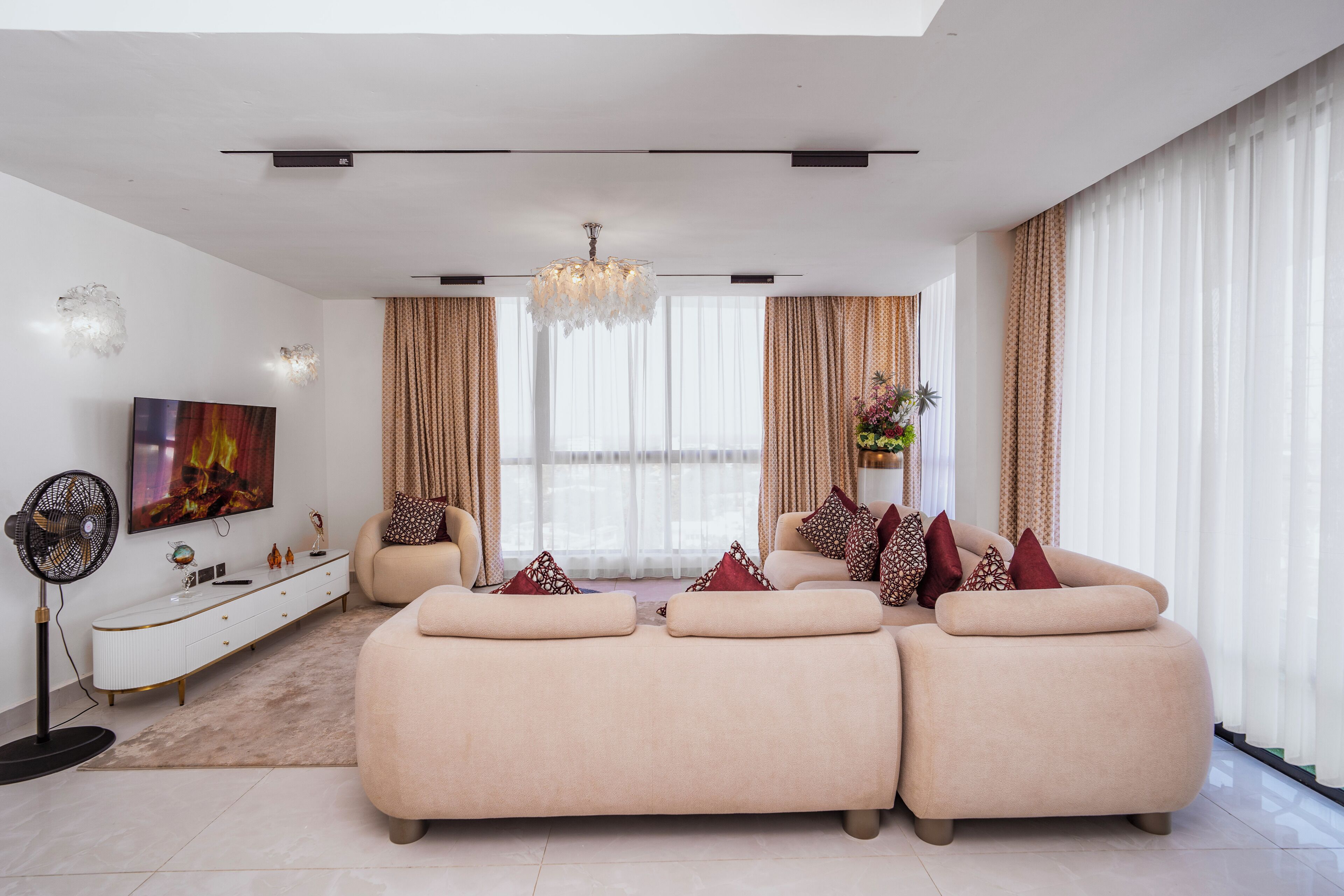 A modern living room with a sectional sofa, a flat-screen TV, a decorative fan, and a chandelier.