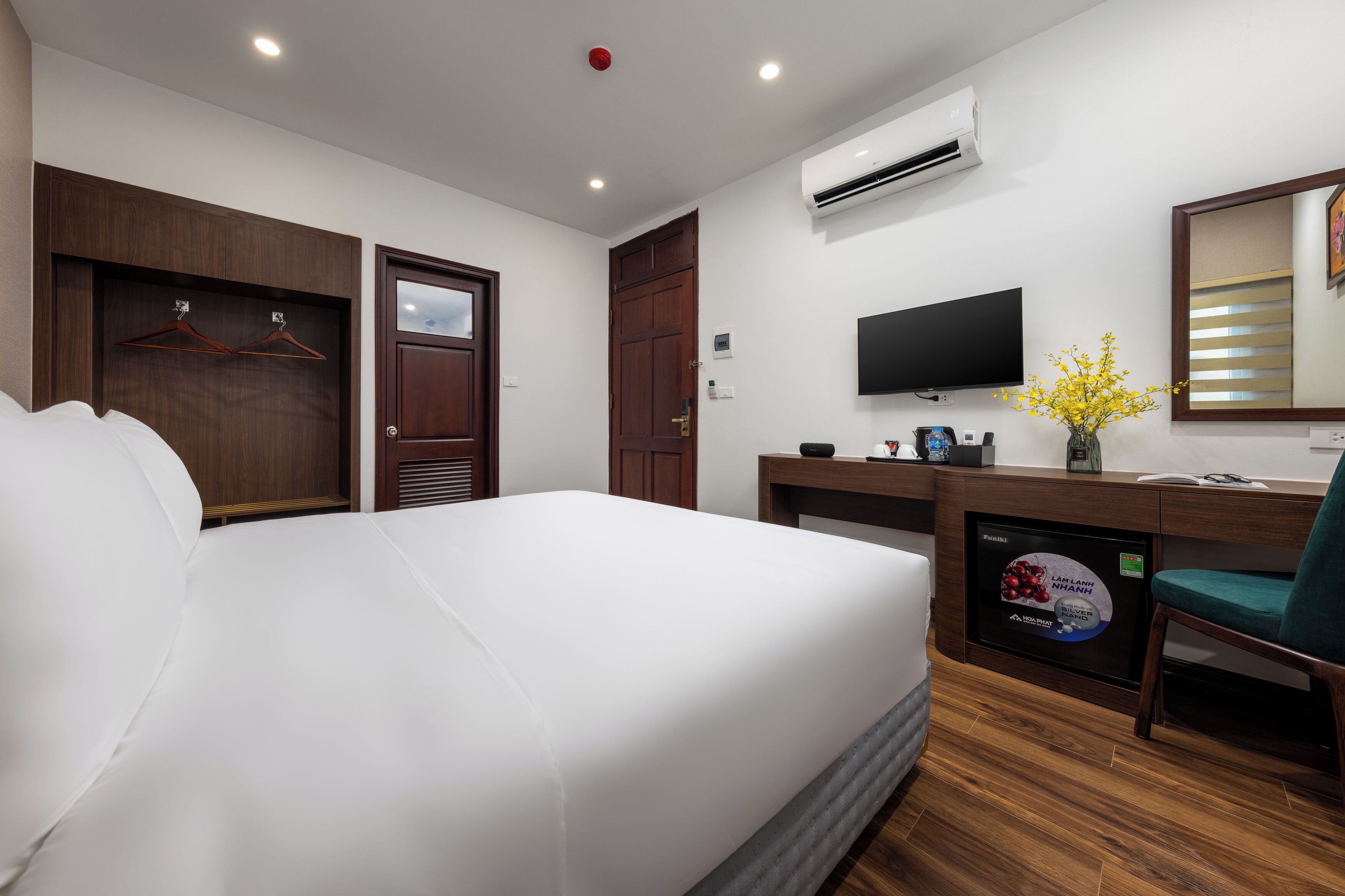 A hotel room with a large bed, a desk with a flat-screen TV, a mirror, and a wall-mounted air conditioner.