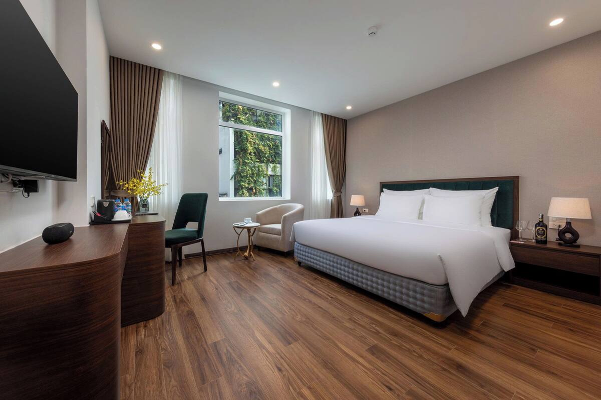 Ein Hotelzimmer mit einem großen Bett, einem Schreibtisch mit Flachbildfernseher, einem Sessel, einem kleinen Tisch und einem Fenster mit Vorhängen.