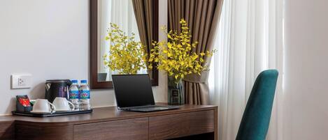 A hotel room with a desk, laptop, coffee maker, water bottles, and a vase with yellow flowers.