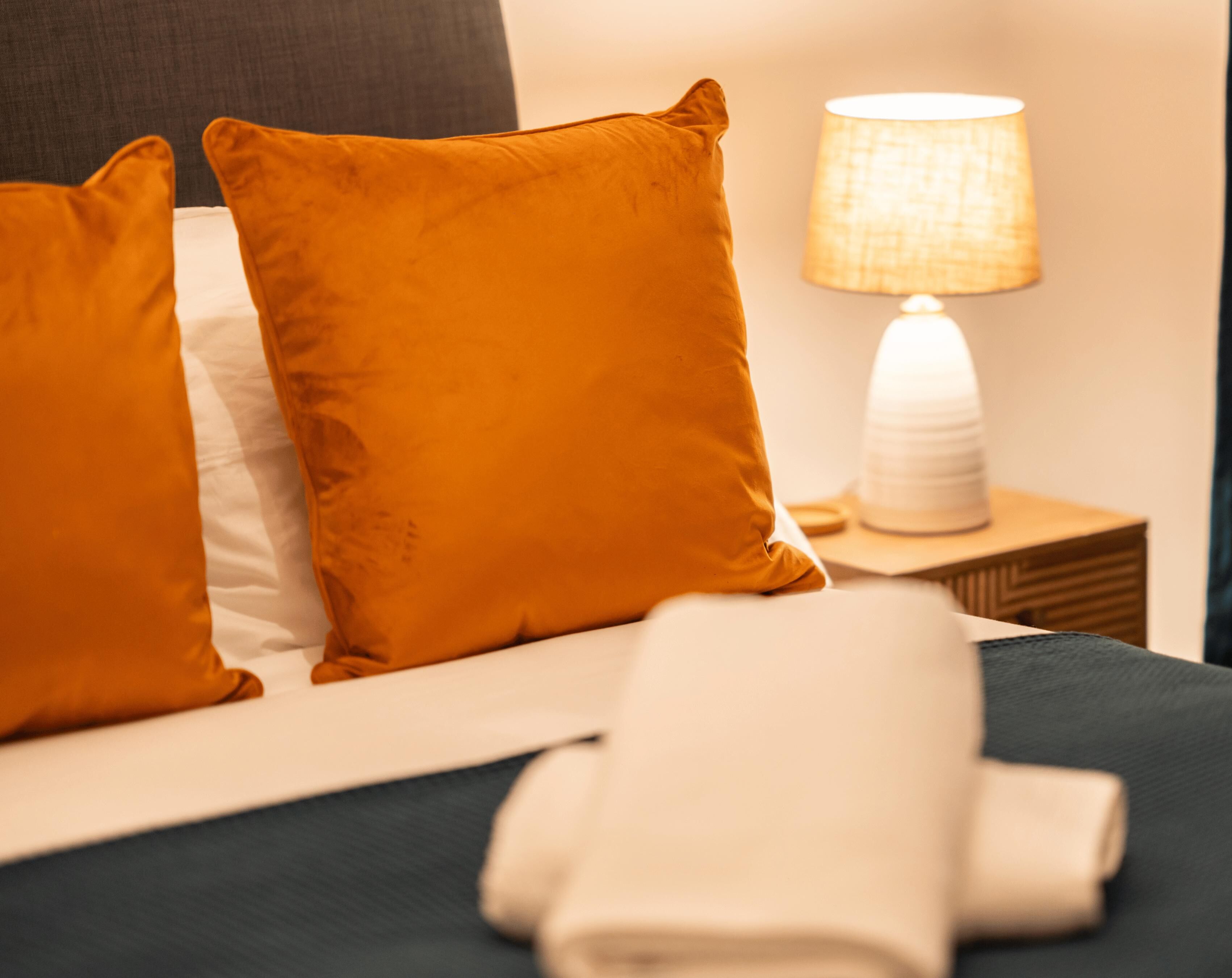 A neatly made bed with two orange pillows and a white towel, a bedside lamp, and a wooden nightstand.