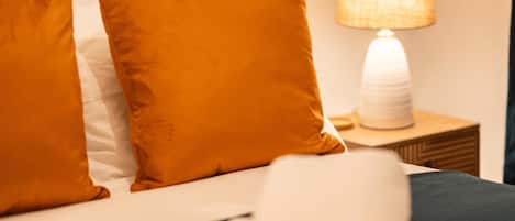 A neatly made bed with two orange pillows and a white towel, a bedside lamp, and a wooden nightstand.