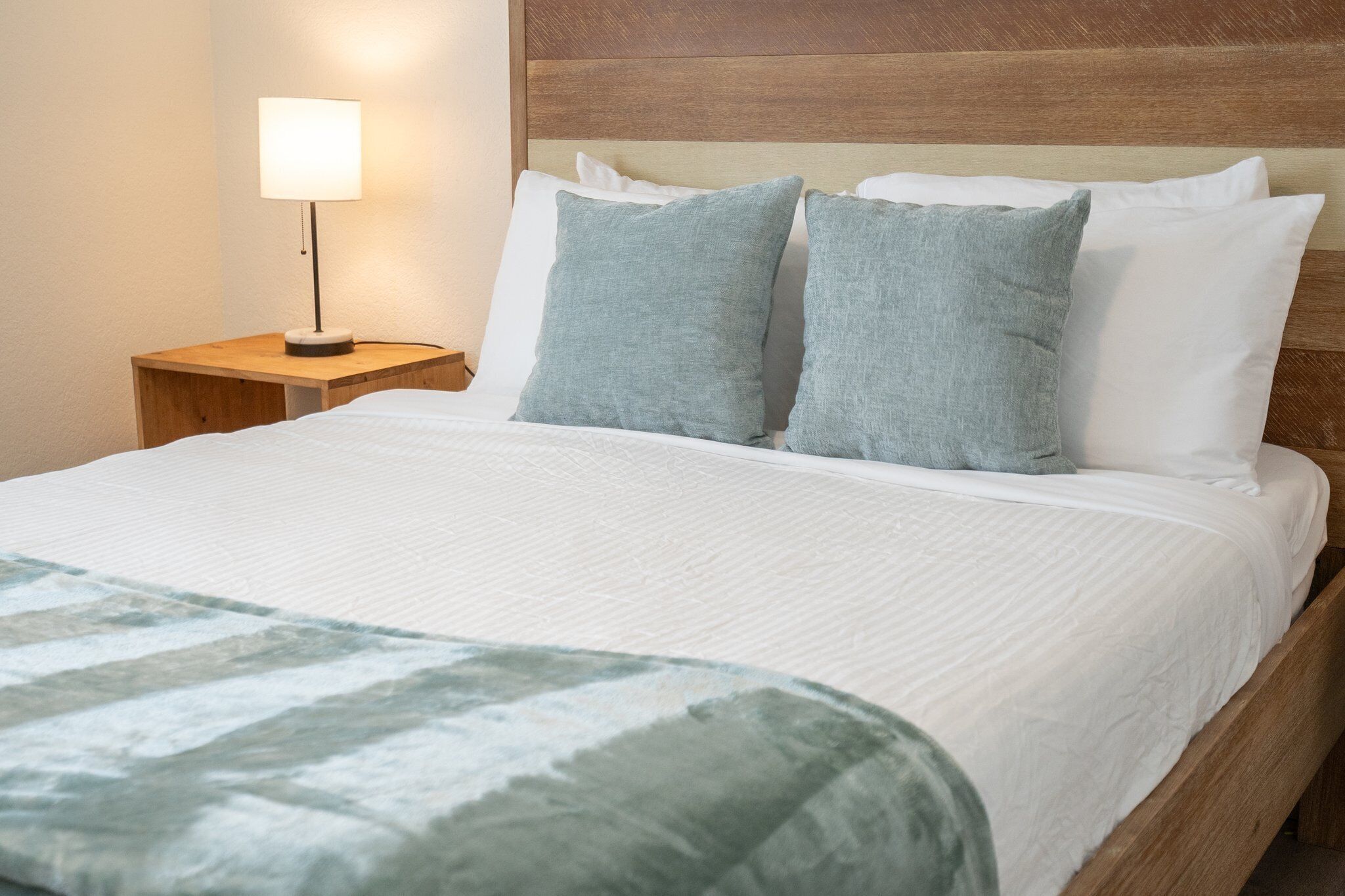 A neatly made bed with a white comforter and two pillows, a bedside lamp, and a wooden headboard.