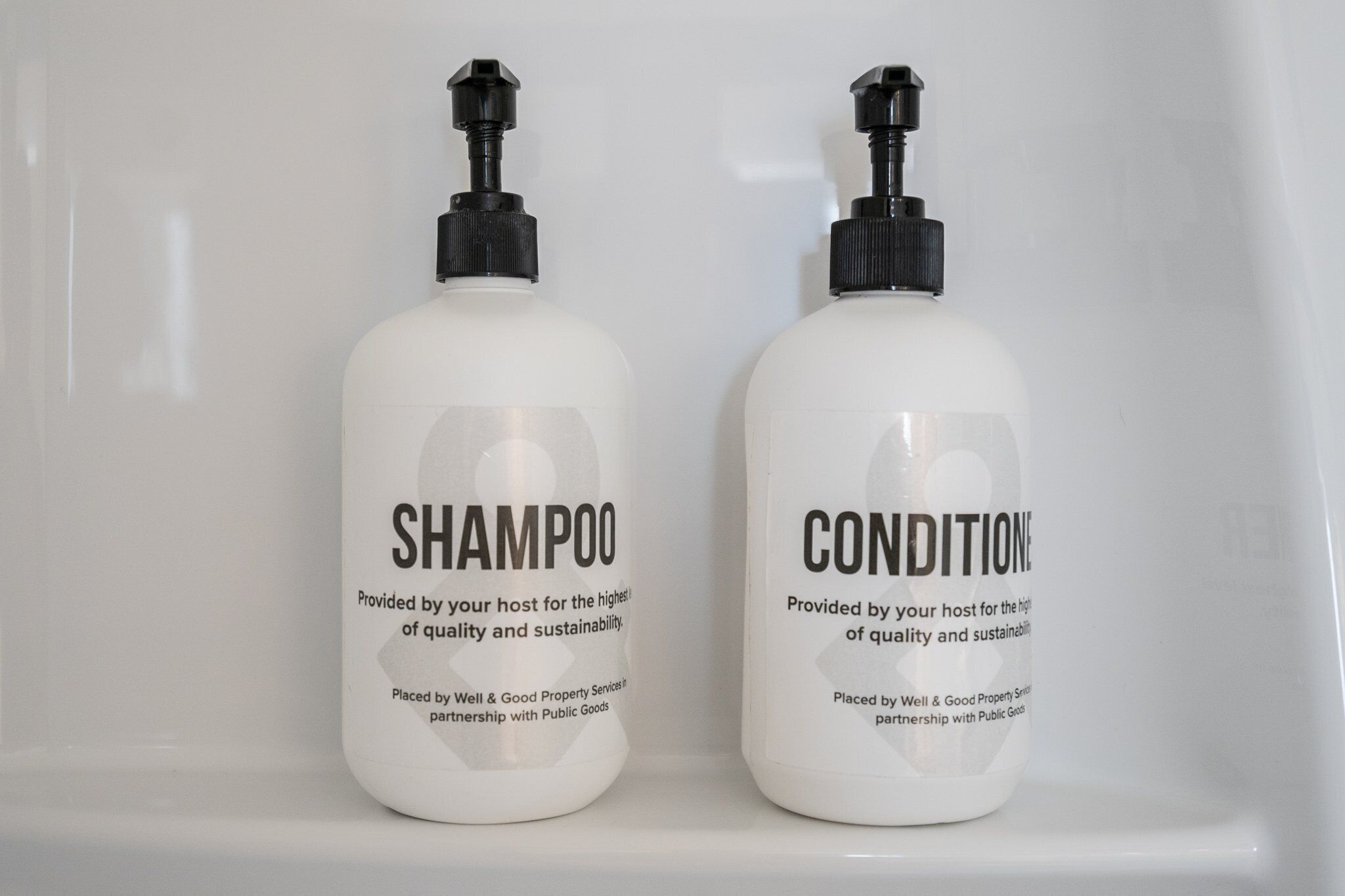 Twee witte flessen met de tekst 'SHAMPOO' en 'CONDITIONER' op een witte plank.