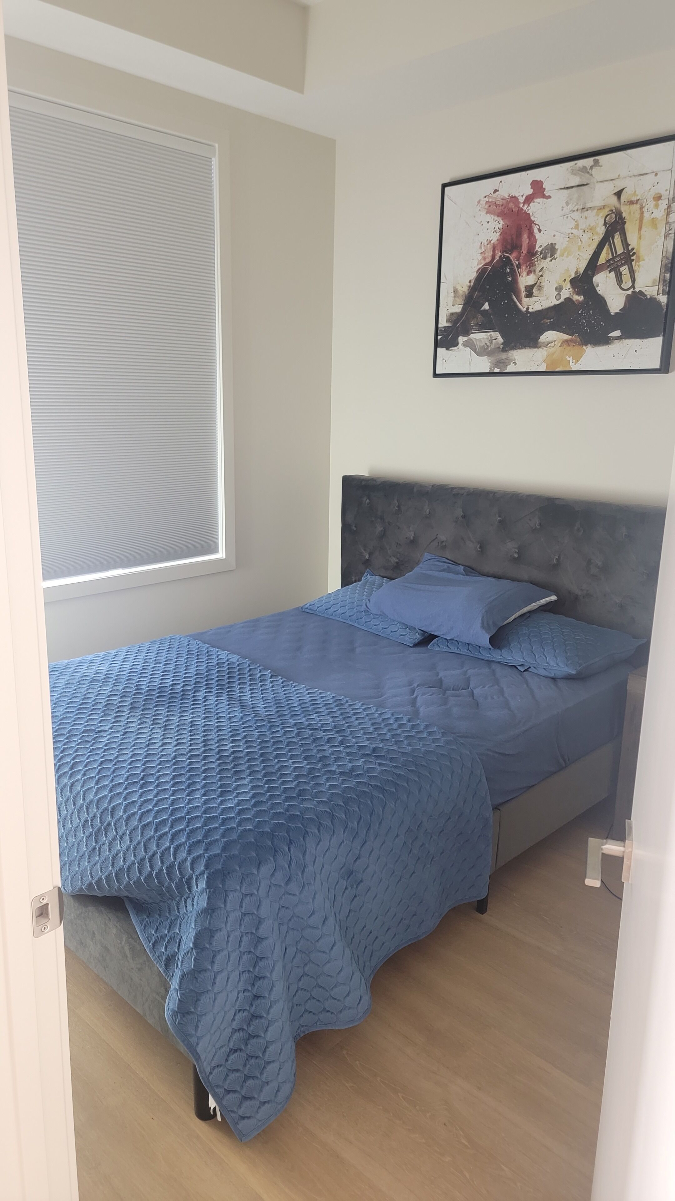 A bedroom with a bed, a window with blinds, a painting on the wall, and a wooden floor.