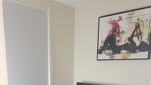 A bedroom with a bed, a window with blinds, a painting on the wall, and a wooden floor.