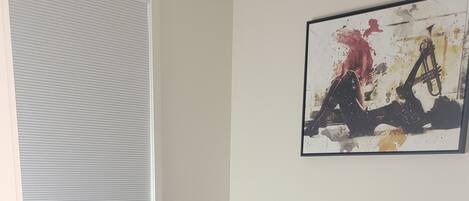 A bedroom with a bed, a window with blinds, a painting on the wall, and a wooden floor.