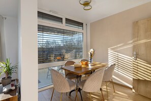 Un comedor con mesa de madera, cuatro sillas, un florero con flores y vistas a través de una ventana con persianas.