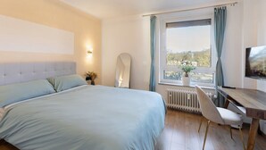 Un dormitori amb llit, escriptori, cadira, radiador, finestra amb cortines i vista exterior.