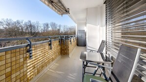 Un balcó amb vista a arbres, una cadira, un taulell i una finestra amb persianes.