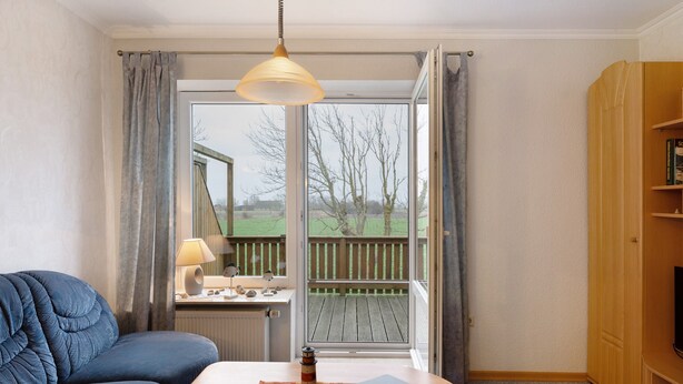 Une pièce avec un canapé bleu, une table en bois sur laquelle se trouve une lampe, un ordinateur portable, et une vue sur une terrasse en bois à l’extérieur.