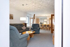 Een woonkamer met een bank, fauteuil en salontafel, een eethoek met tafel en stoelen, en een keuken op de achtergrond.