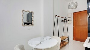 Una stanza piccola e pulita con un tavolino rotondo bianco, due sedie bianche, uno specchio con cornice ondulata, una porta in legno e una lampada a sospensione.