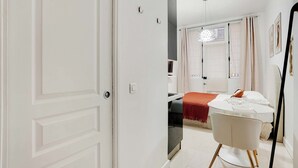 Una camera d'albergo moderna con un letto, un tavolo da pranzo e una sedia. Si nota una porta a sinistra e uno specchio a destra.