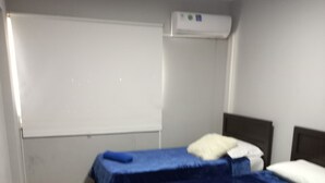 Ein Zimmer mit zwei Betten, jeweils mit blauer Decke und Kissen, einem kleinen Tisch und einer Klimaanlage an der Wand.
