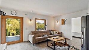 Een moderne woonkamer met een bank, een salontafel, een eettafel, een koelkast en uitzicht op groen.
