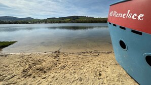 Een boot met 'MS Diemelsee' op de zijkant, een zandstrand, kalm water en heuvels in de verte.