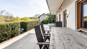 Een terras met buitenmeubilair, een haag en een huis op de achtergrond.