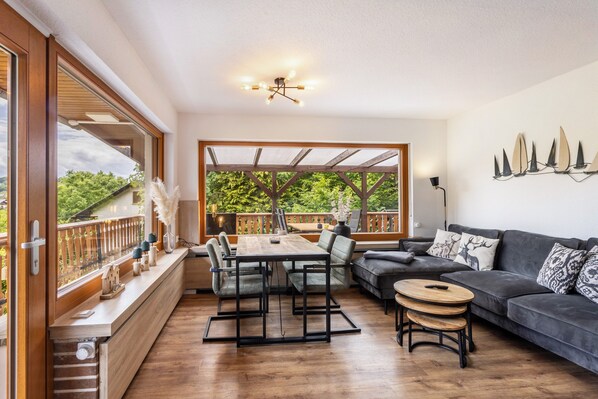 A spacious living area with a wooden dining table, a sofa, and a balcony with a view of greenery.