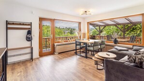 A spacious living area with a wooden floor, a sofa, a dining table with chairs, and a balcony with a view of greenery.