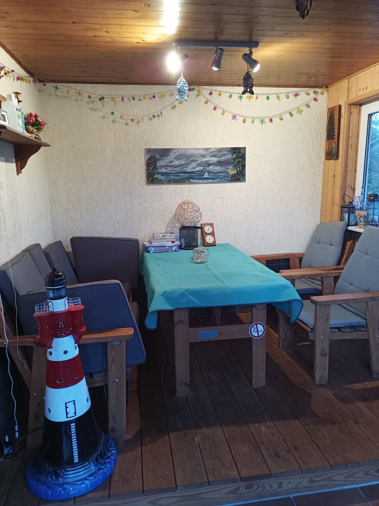 A cozy room with a wooden ceiling, a table with a green tablecloth, two chairs, a bookshelf, a painting, and a decorative lighthouse.