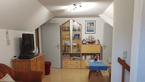 Un salon chaleureux avec un téléviseur, un meuble en bois, une bibliothèque et un coin repas comprenant une table et des chaises.