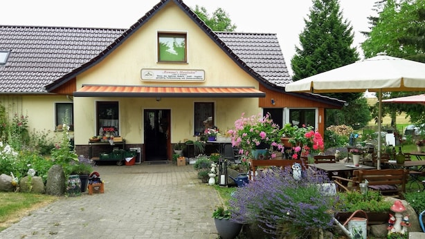 Un bâtiment pittoresque avec un toit en tuiles, une enseigne et un jardin agrémenté de plantes en pot et d’un espace aménagé pour s’asseoir à l’extérieur.