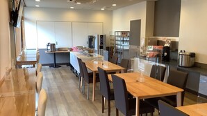 A modern dining area with wooden tables and chairs, a kitchen area with appliances, and a view of the city. - Smile Hotel Yonezawa (Yonezawa)