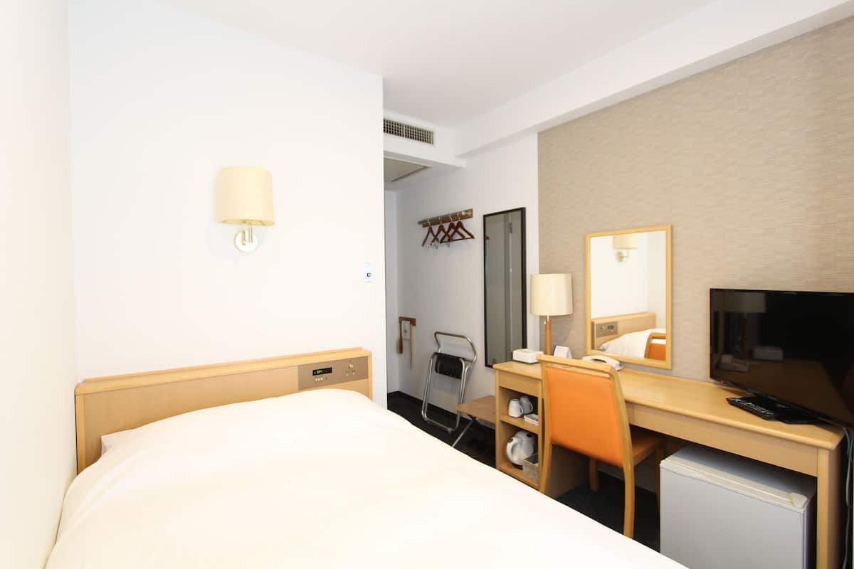 A hotel room with a bed, a desk, a chair, a small refrigerator, a mirror, and a wall-mounted lamp.