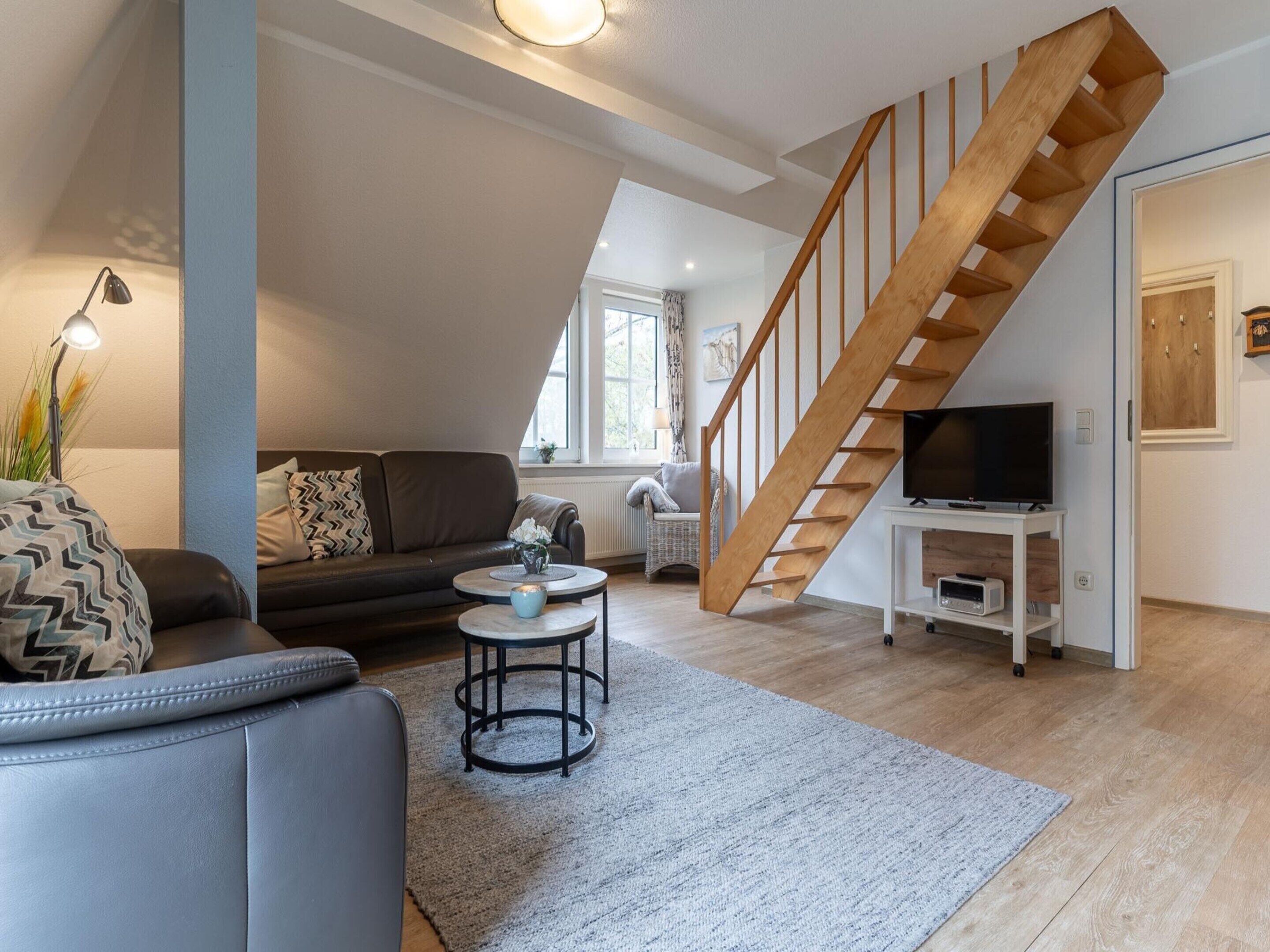 A modern living room with a sofa, coffee table, and a TV on a stand. A wooden staircase is visible in the background.
