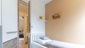 Un dormitorio con una cama individual, cabecera blanca, una pequeña estantería sobre la cama y una puerta que da a otra habitación.