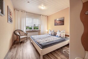 Ein Schlafzimmer mit Bett, Sessel, Fenster mit Jalousien und einem Wandbild.