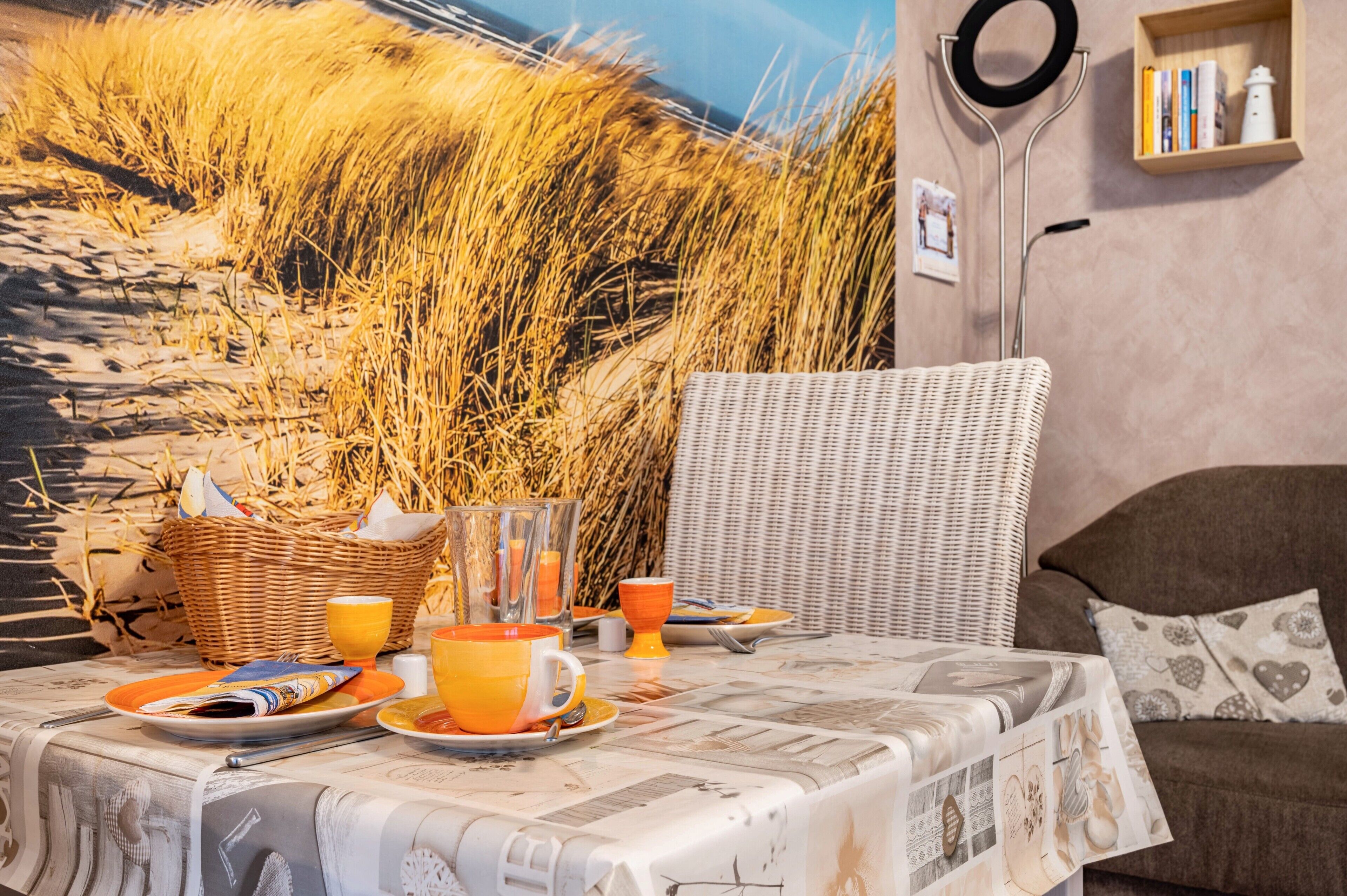 A breakfast table set with orange plates, cups, and a wicker basket, a wicker chair, and a bookshelf with books and a kettle.