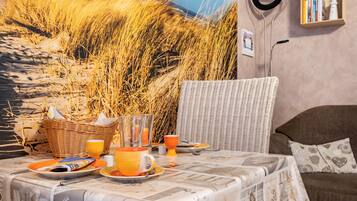 A breakfast table set with orange plates, cups, and a wicker basket, a wicker chair, and a bookshelf with books and a kettle.