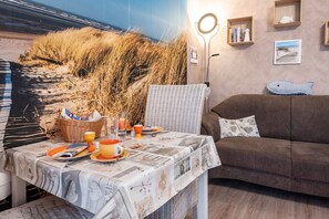 Una habitación con una mesa de comedor preparada para dos personas, una silla de mimbre, un sofá marrón y una pared decorada con un mural de playa.