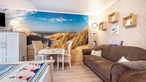 Ein Zimmer mit Wandgemälde vom Strand, einem braunen Sofa, einem Esstisch mit Stühlen und einem weißen Schrank.