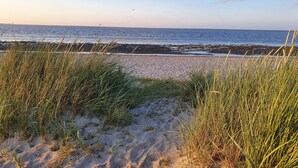 Ein Strand mit Sand und Gras im Vordergrund, einem klaren Himmel und dem Meer im Hintergrund.