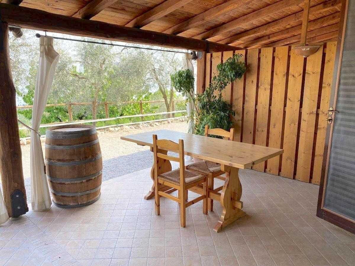Et træbord og stole under en overdækket terrasse med grønne omgivelser i baggrunden.