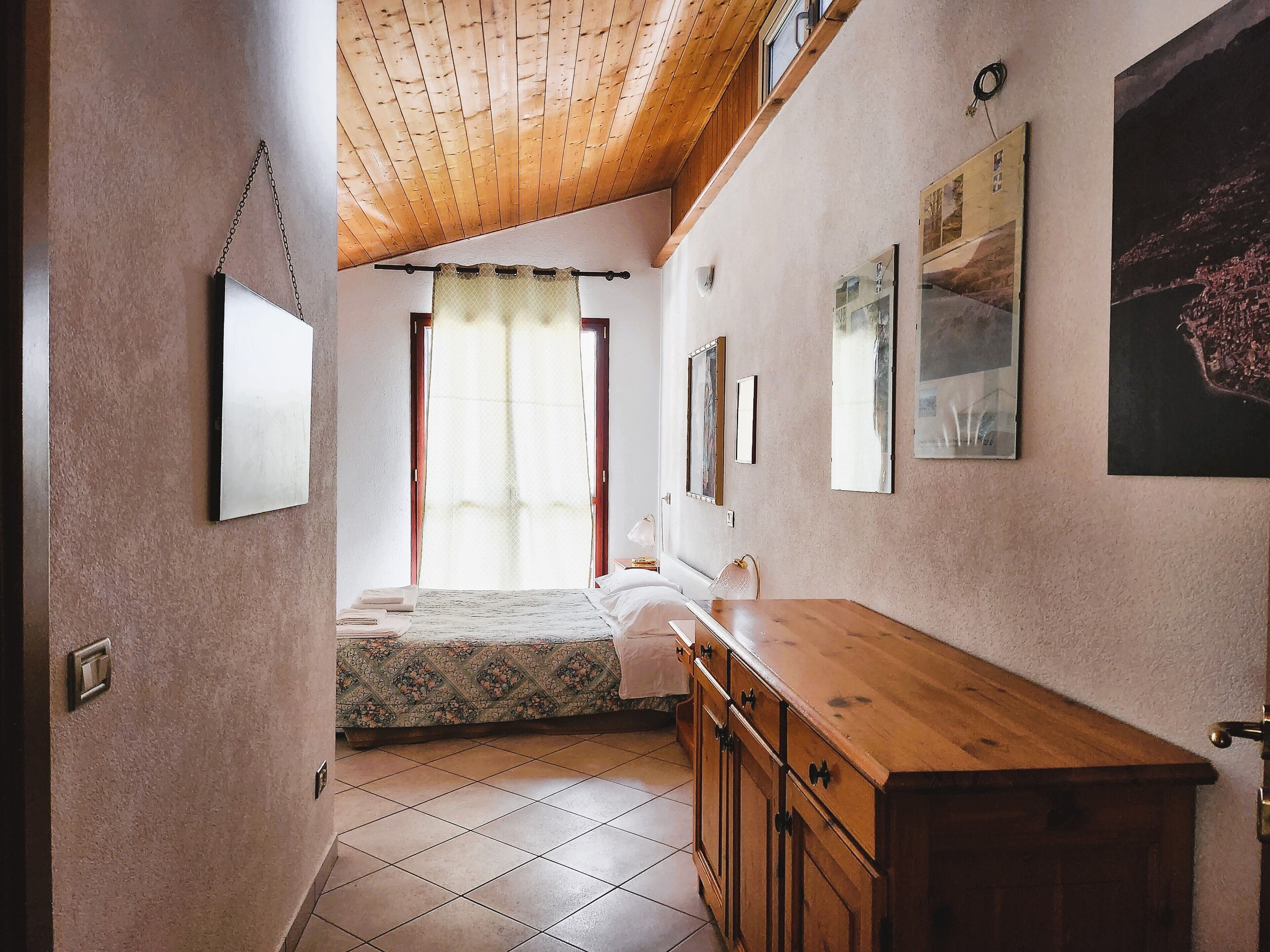 Une chambre à coucher avec un lit, une commode en bois, une fenêtre avec des rideaux et des tableaux accrochés au mur.